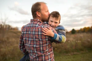 Father hugging smiling son in a field during sunset – symbol of love, security, and family resilience.
